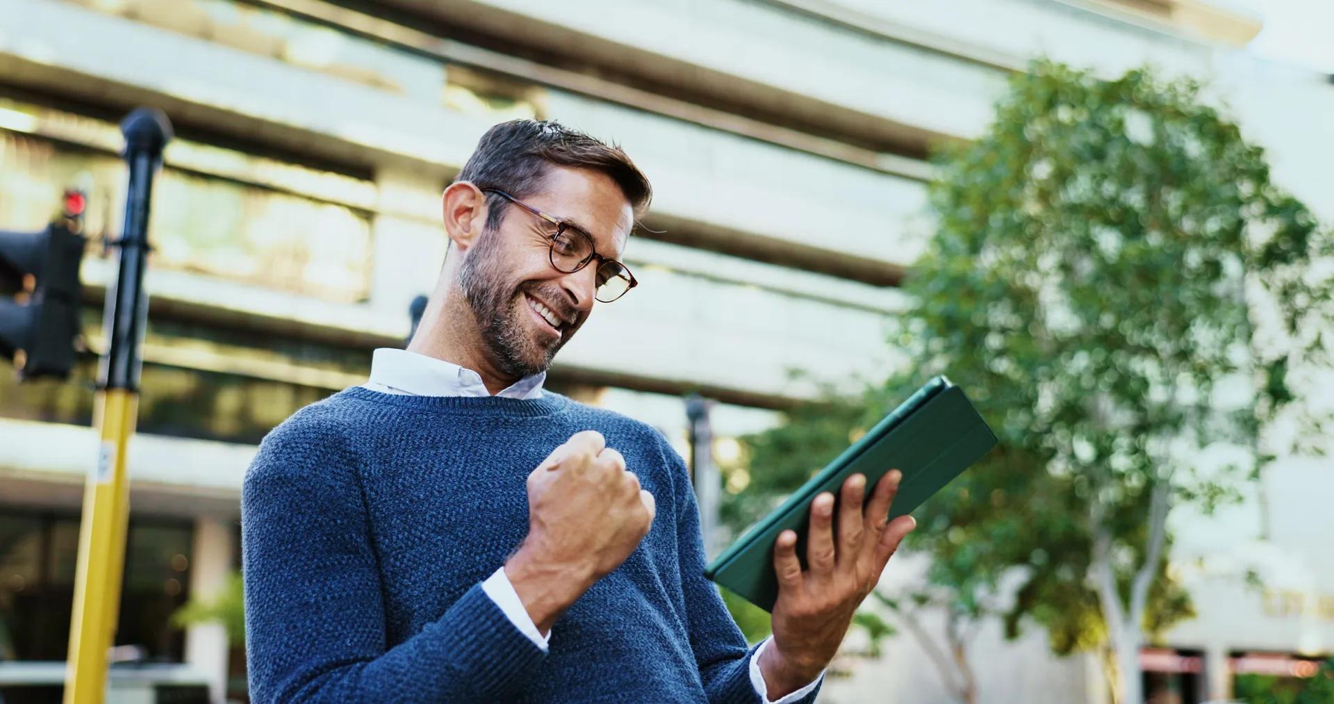 Homem comemorando ao consultar um tablet em frente a um edifício