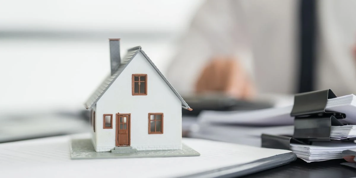 Uma maquete de uma casa branca sobre uma mesa e ao fundo um homem de camisa e gravata sentado