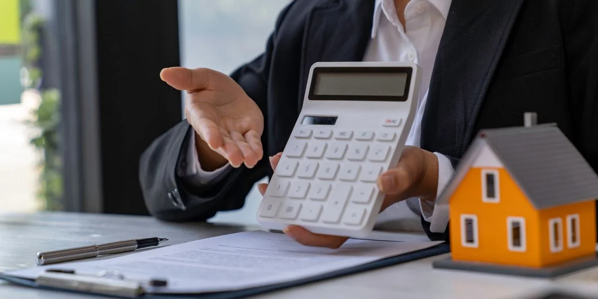 Uma pessoa apresentando uma calculadora sobre uma mesa com um caderno e uma miniatura de uma casa